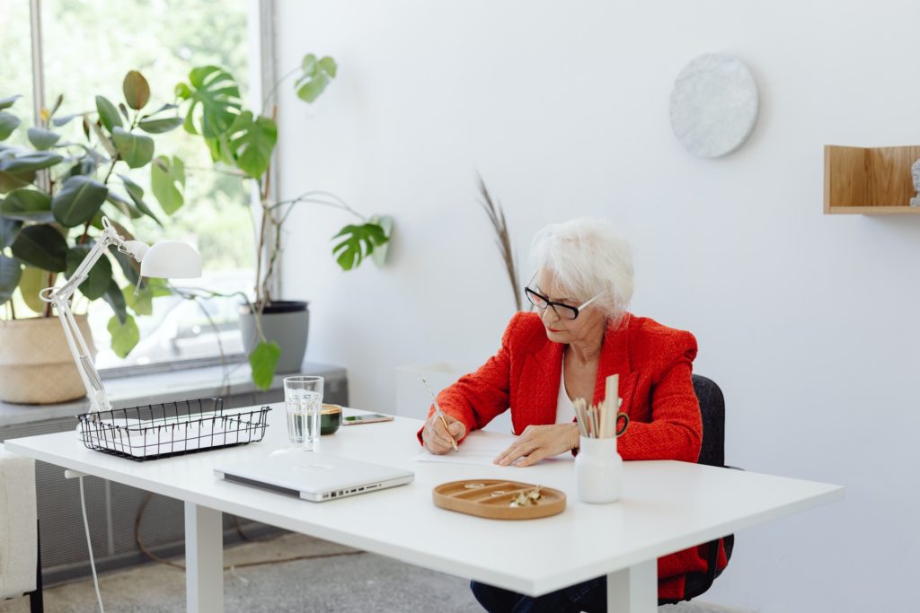 Mulher de cabelos grisalhos e casaco vermelho está sendada em mesa de trabalho e assina documentos