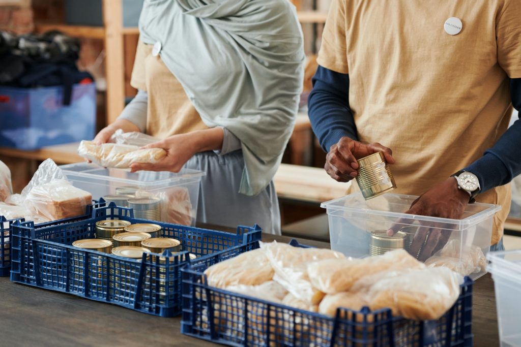 Sobre uma mesa, voluntários, uma mulher branca e um homem negro, organizam latas de alimentos e pães em caixas de plástico para transporte