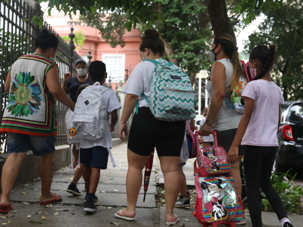 adultas acompanham crianças com uniformes escolares e mochilas a caminho da escola