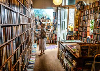 Mulher branca, de cabelos grisalhos, está parada no corredor de uma livraria e olha com atenção para as prateleiras. Ela está de perfil, no centro da imagem