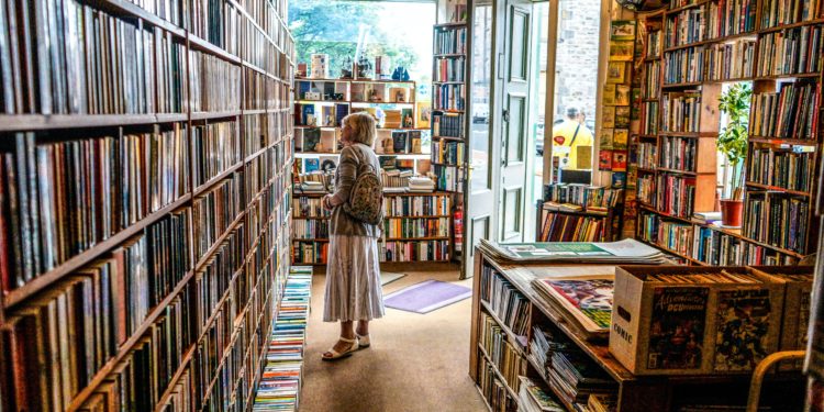 Mulher branca, de cabelos grisalhos, está parada no corredor de uma livraria e olha com atenção para as prateleiras. Ela está de perfil, no centro da imagem