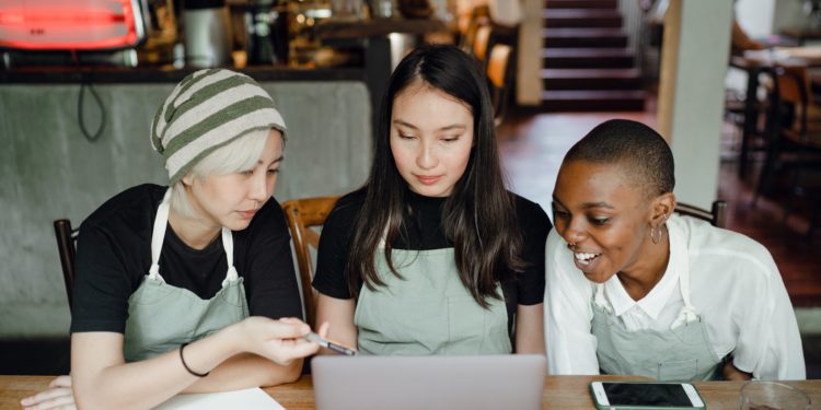 De frente para a câmera,rês mulheres estão sentadas em uma mesa e olham para a tela de um computador. Elas estão em um estabelecimento semelhante a um café ou restaurante. Uma das mulheres é asiática, tem cabelos loiros e usa gorro, a do meio tem pele branca e cabelos castanhos longos, e a da direita é negra e tem cabelo raspado