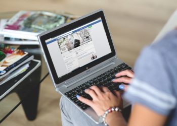 Mulher utiliza notebook apoiado no colo e acessa página do Facebook. Ela tem unhas pintadas de azul.