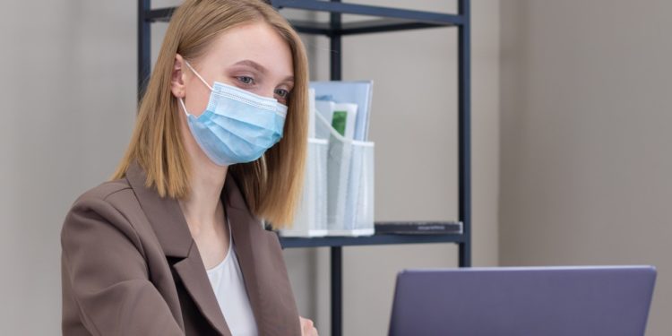 Mulher de cabelos loiros usa notebook em mesa de trabalho. Ela máscara