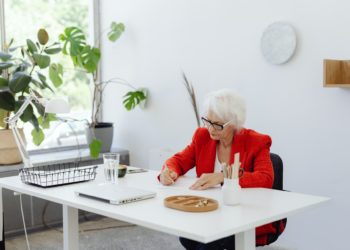 Mulher de cabelos grisalhos e casaco vermelho está sendada em mesa de trabalho e assina documentos; Além do preconceito de gênero, mulheres mais velhas sofrem com a desvalorização de seus corpos, diz pesquisa da FGV EAESP