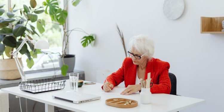Mulher de cabelos grisalhos e casaco vermelho está sendada em mesa de trabalho e assina documentos; Além do preconceito de gênero, mulheres mais velhas sofrem com a desvalorização de seus corpos, diz pesquisa da FGV EAESP