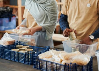 Sobre uma mesa, voluntários, uma mulher branca e um homem negro, organizam latas de alimentos e pães em caixas de plástico para transporte