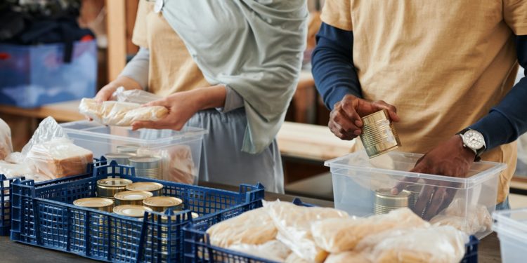 Sobre uma mesa, voluntários, uma mulher branca e um homem negro, organizam latas de alimentos e pães em caixas de plástico para transporte