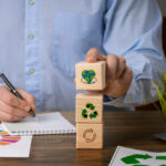 homem escreve em papel enquanto empilha blocos de madeira com ilustrações que representam temas ambientais, como o símbolo da reciclagem e o planeta Terra, remetendo à importância da gestão ambiental nas políticas públicas