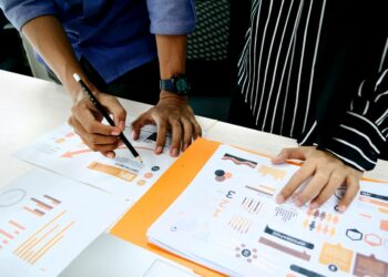 mãos de duas pessoas estão sobre documentos com informações e infográficos dispostos sobre mesa de trabalho