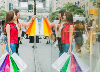 Duas mulheres brancas seguram sacolas coloridas e observam vitrine; Sustentabilidade e ética empresarial contribuem para a percepção do consumidor sobre o valor da marca