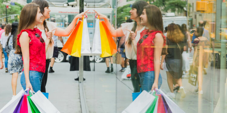 Duas mulheres brancas seguram sacolas coloridas e observam vitrine; Sustentabilidade e ética empresarial contribuem para a percepção do consumidor sobre o valor da marca