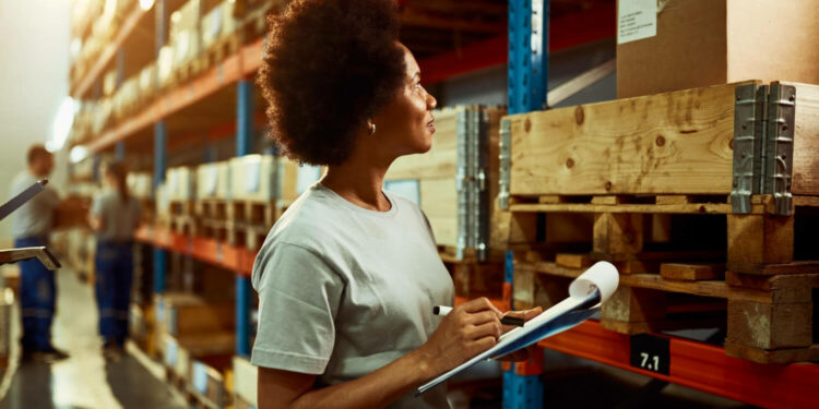 mulher jovem negra observa caixas em estoque e faz anotações em prancheta