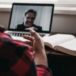pessoa gesticula em frente a tela de computador sobre mesa de trabalho; a tela mostra imagem de homem branco de meia idade em videoconferência