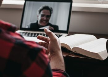 pessoa gesticula em frente a tela de computador sobre mesa de trabalho; a tela mostra imagem de homem branco de meia idade em videoconferência
