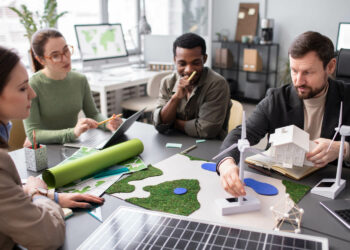 Quatro pessoas, duas mulheres brancas, um homem negro e um homem branco, sentados ao redor de mesa, observam modelos de turbinas de energia eólica e painéis solares