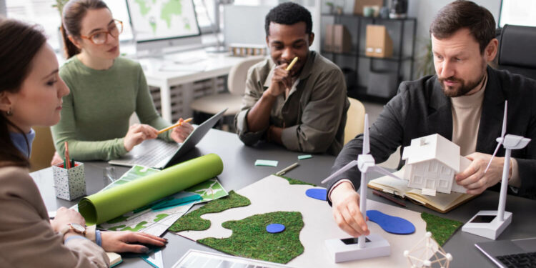 Quatro pessoas, duas mulheres brancas, um homem negro e um homem branco, sentados ao redor de mesa, observam modelos de turbinas de energia eólica e painéis solares